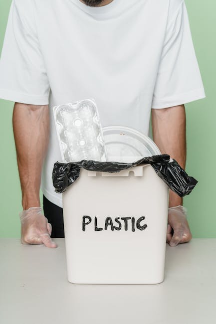 A person places a crumpled brown paper bag into a grey rubbish bin, which is positioned on a wooden surface indoors. The bin has a partially visible lid and appears to be designated for general waste