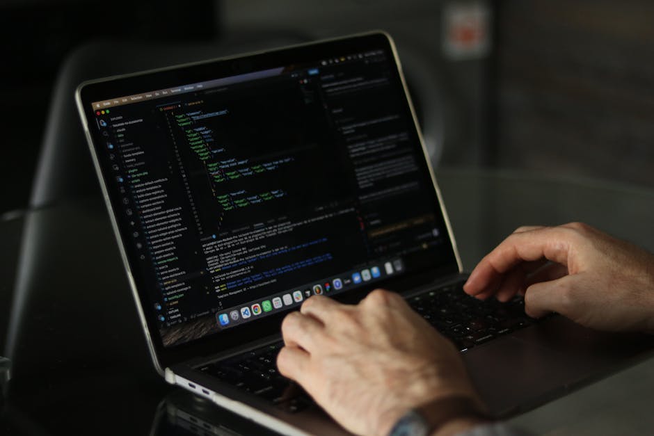 A person wearing a light blue shirt is sitting at a desk, working on a laptop with an open code editor displaying multicolored lines of programming code. The individual is holding a pen in their right